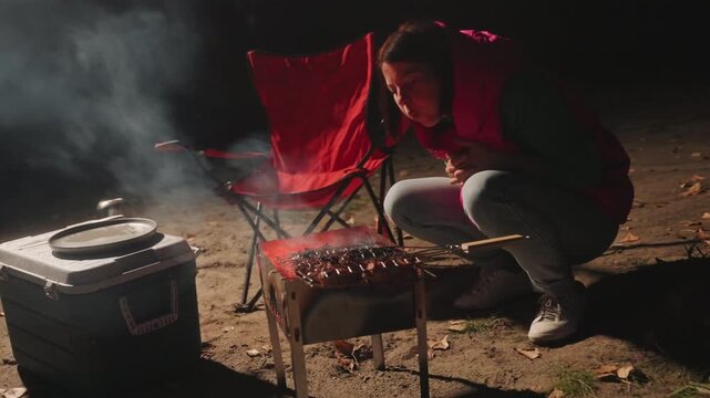 Camping night grilling camper crouches by portable grill, cooler and red folding chair nearby, smoke rising from glowing coals, hands tending embers, warm glow illuminating shore, focused campsite