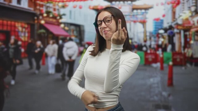 Young hispanic woman in white crop top and glasses shows middle finger and beckoning motion with bare finger and slight smirk in a crowded street market lined with lanterns; defiance.