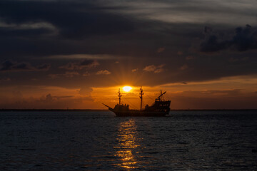A sailing ship is silhouetted against a vibrant sunset, casting a warm glow over the ocean. The sky's hues range from deep orange to soft pink, reflecting beautifully on the water's surface. © dfriend150