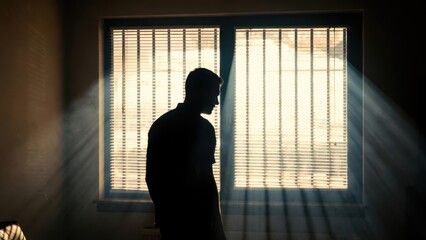 Fototapeta premium Man standing in a dimly lit room, with light rays streaming through the window, evoking themes of isolation, introspection, and dramatic storytelling for film and emotional content marketing.