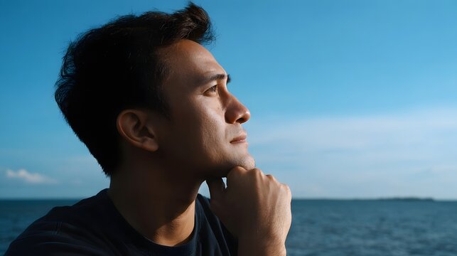 A thoughtful man rests his chin on his hand while gazing out over the vast blue ocean horizon under a clear sky