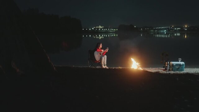 Lakeshore night white camper rehearsing guitar, camping gear and portable amplifier near small fire, skyline lights across water, intimate practice session, cool breeze and smoke, solo performer