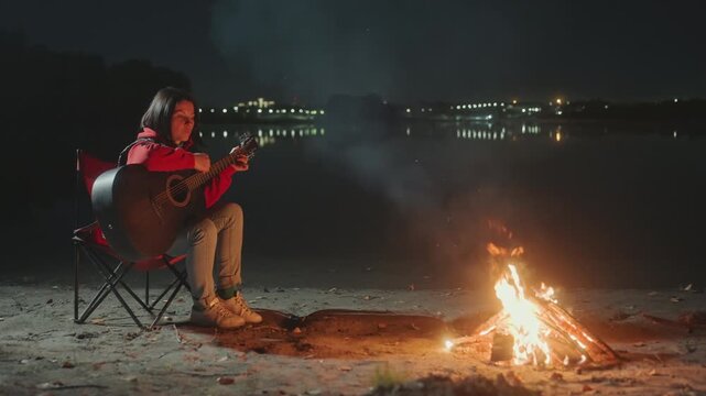 Lakeside night camping with guitar, caucasian woman sits in red folding chair by bonfire, strumming acoustic guitar beside calm lake, glowing embers and soft smoke, distant city lights reflecting