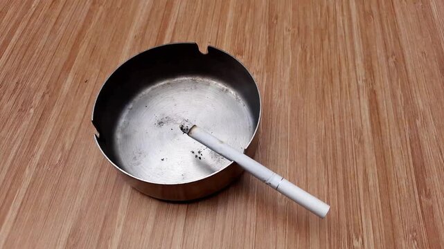 Lit cigarette rests inside a metal ashtray on a wooden surface as wisps of smoke rise in this slow motion video clip depicting a dangerous daily habit.