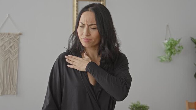 Young hispanic woman rubbing and clutching her shoulder, wincing and shifting posture in a home studio with macrame wall and plants; pain recovery.