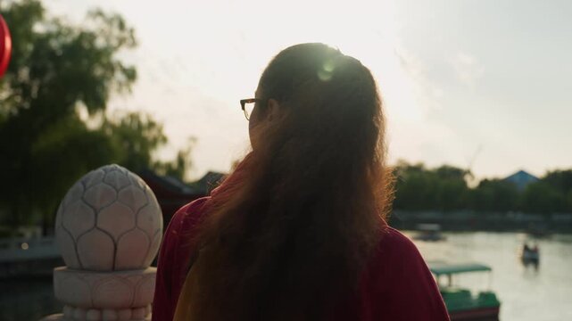 Young asian woman by river, student leaning on stone balustrade, observer of passing boats, traveler with backpack, thinker facing golden light, soft backlight on curly ponytail, glasses catching sun,