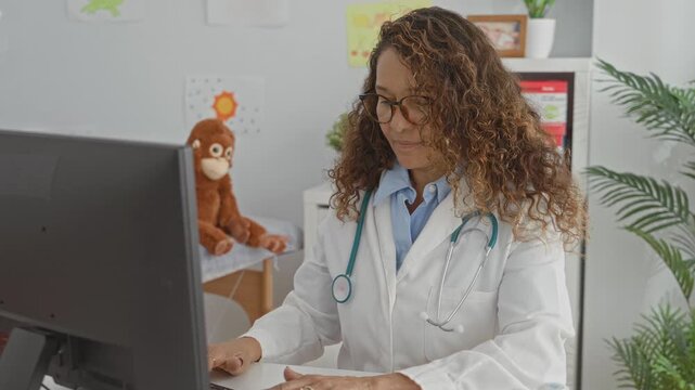 Woman doctor types on keyboard at computer with stethoscope and glasses, seated near children's drawings and toy in clinic building; focused compassionate care.