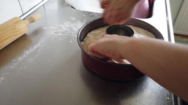 Hands pressing and shaping raw dough into a ring cake pan on a floured stainless countertop, conveying home baking, preparation, texture and the comforting ritual of making homemade bread or cake.