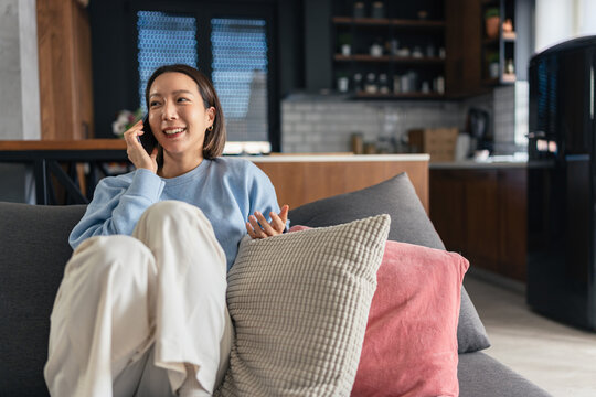 Smiling Asian woman talking on smartphone while relaxing on a sofa at home. Casual lifestyle scene showing communication, comfort and modern everyday living indoors