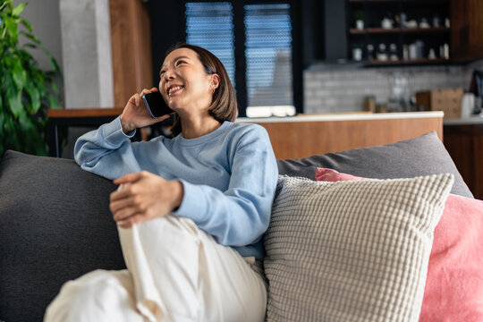 Smiling Asian woman talking on smartphone while relaxing on a sofa at home. Casual lifestyle scene showing communication, comfort and modern everyday living indoors