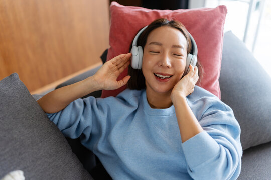 East Asian woman relaxing on a sofa at home, smiling while holding headphones and enjoying music. Cozy, joyful moment of leisure and relaxation in a modern living space.