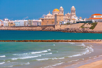 Wide coastal view of Cadiz, Spain, with sandy beach, turquoise Atlantic water, breakwaters, and the cathedral skyline under clear daylight.