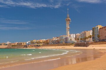 Sunny coastal view of La Caleta beach in Cadiz, Spain, with sandy shore, gentle Atlantic waves, city skyline, and Tavira tower in the background.