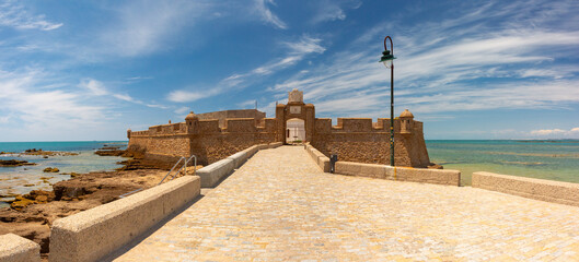 Panoramic coastal view of San Sebastian fortress in Cadiz, Spain, with stone entrance causeway, rocky shoreline, and calm Atlantic water under clear blue sky.