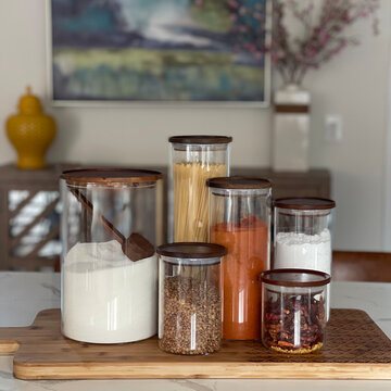 A clean and aesthetically pleasing shot of four glass canisters filled with kitchen essentials like pasta, flour, and grains. The jars feature elegant wooden lids and are arranged on a bamboo surface.