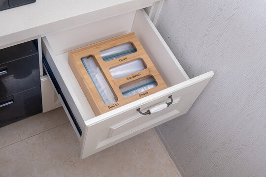 A high-angle shot of an open white kitchen drawer featuring a wooden bamboo organizer for food storage bags. The organizer has labeled slots for Gallon, Quart, Sandwich, and Snack bags