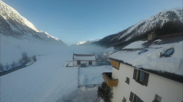 Snow-covered mountains embrace a serene valley, where quaint buildings nestle amidst a blanket of white, creating a peaceful and picturesque winter landscape.
