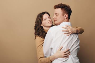 Fototapeta premium Young couple embraces tenderly against beige background. Woman in brown sweater hugs man in white shirt. Concept of love, affection, intimacy, and relationship between partners.