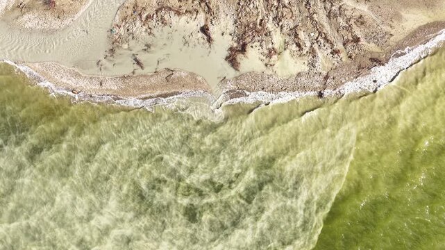 An aerial drone shot of the natural shoreline where waves crash against the shore and meet the muddy water. A bird&rsquo;s-eye view showing erosion and the movement of water.