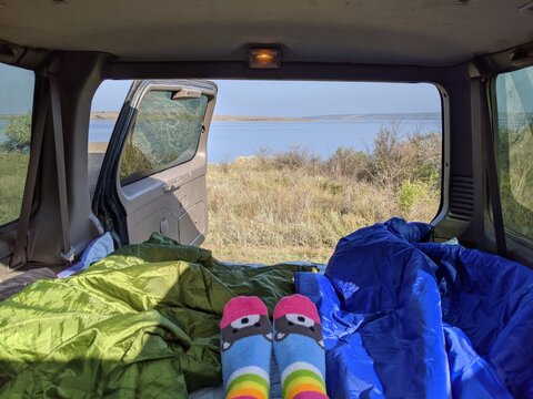 Cozy POV of waking up in a car during a road trip, view from the trunk on a peaceful lake and autumn landscape, colorful socks and sleeping bags, van life and camping aesthetic