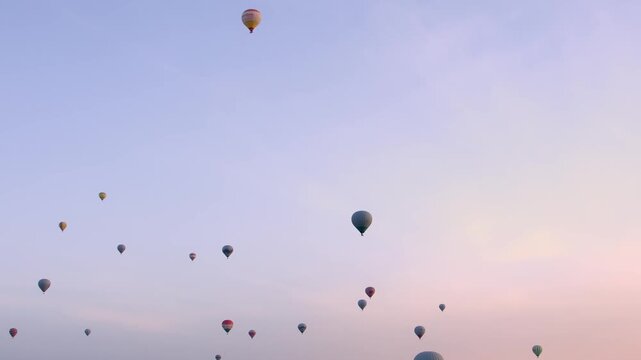 A scenic wide shot of numerous colorful hot air balloons floating gently in the clear, pastel-colored morning sky over the unique rock formations and valleys of Cappadocia, Turkey. This iconic tourist