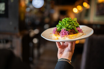 Obraz na płótnie Canvas A waiter holds a white plate with a gourmet open-faced lamb burger topped with pickled red onions and fresh greens in a dimly lit, upscale restaurant or bar setting.