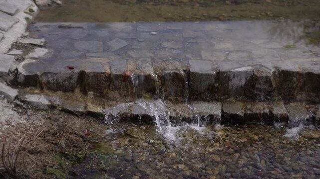 Water flows over a small stone ledge forming a miniature waterfall in a shallow pond