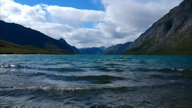 A pristine lake reflects the sky, framed by majestic mountains under a partly cloudy sky, creating a serene and picturesque natural landscape.