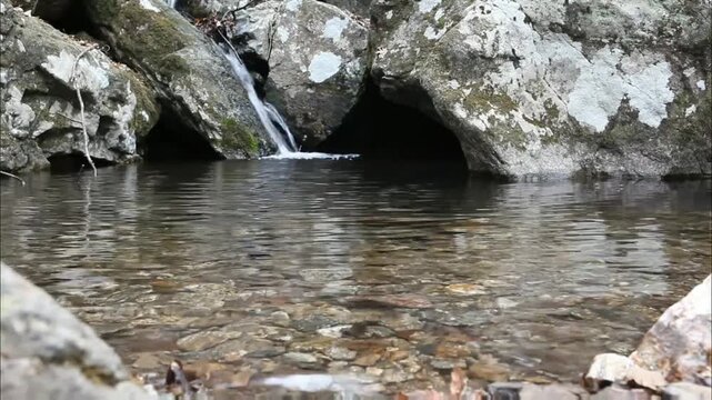 A small waterfall cascades into a clear pool, surrounded by mossy rocks. The water reflects the overcast sky, creating a peaceful, natural scene.