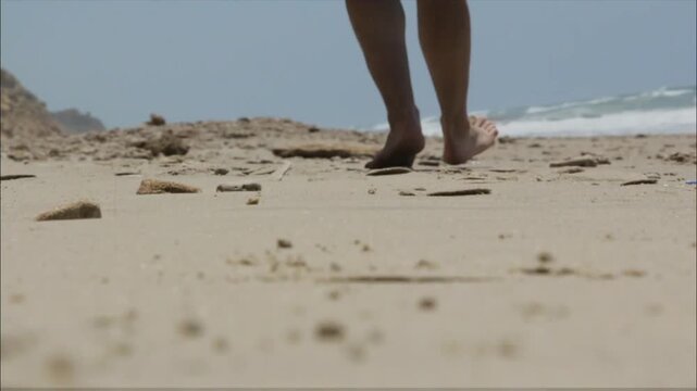 A person walks barefoot on the beach, their feet sinking into the sand as they approach the ocean under a bright sky with distant waves.