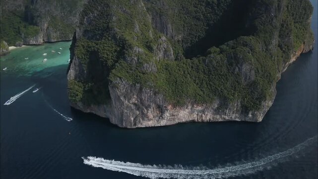 Towering, verdant cliffs meet the turquoise sea, boats leaving white trails on the water's surface, showcasing a dramatic and beautiful tropical island paradise.
