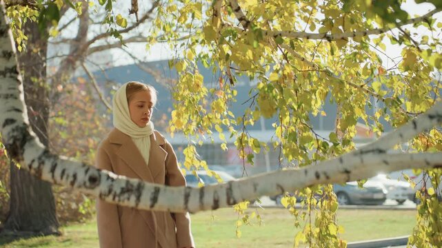 White woman walking beneath birch branches in urban park, wide shot of commuterobserver pausing near trees with cars and buildings in background beige coat and scarf, autumn leaves drifting, sunlight