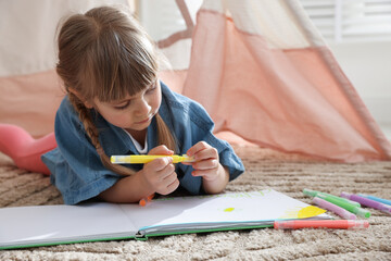 Little girl drawing in sketchbook near toy wigwam at home © New Africa