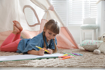 Little girl drawing in sketchbook near toy wigwam at home © New Africa