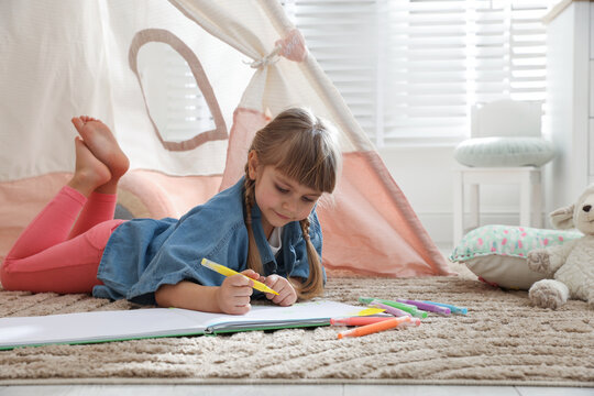 Little girl drawing in sketchbook near toy wigwam at home