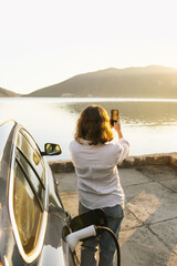 Fototapeta premium Woman with smartphone next to charging electric car on the seacost during summer vacation