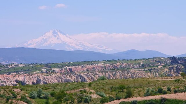A wide, daytime shot showcasing the stunning, otherworldly landscape of Cappadocia in Central Anatolia, Turkey. The foreground features green fields and unique, eroded rock formations, while the backg