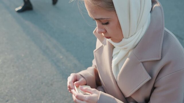 Young woman holding red rose bench, removing petals and counting, wrapped in scarf and coat, empty sidewalk backdrop, golden hour glow, tender yet melancholic atmosphere, subtle cinematic tones.