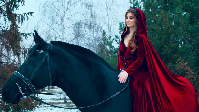 Beautiful noble fantasy medieval woman stroking neck sitting astride black horse. steed hair shines, shimmers under princess hand. Girl smiling happily, red vintage long dress cloak cape head hood.