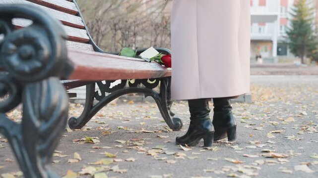 Rose on bench, woman walking away, autumn park closeup of wet wooden seat and single red bloom, long coat and high boots leaving fallen leaves on pathway, soft overcast light, cinematic shallow depth,