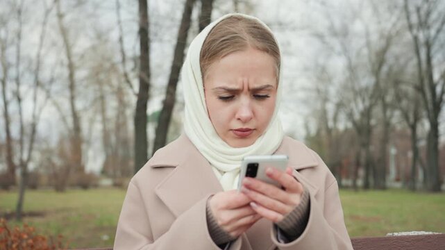 Young white woman looking frustrated furrowed brow and tapping touchscreen, frowning at notification, troubleshooting app or message, beige coat and knit scarf, park trees and overcast sky, candid