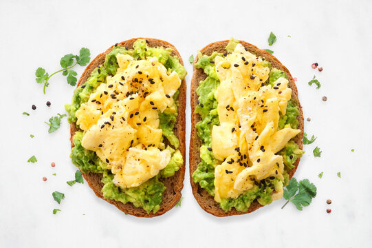 Healthy avocado toast with scrambled eggs on sourdough bread. Top down view on a white marble background.