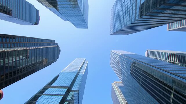 Extreme low-angle video of towering, hyper-modern reflective glass skyscrapers reflecting a clear azure sky with sun flare. A smooth crane and pan emphasizes awe-inspiring scale and aspirational