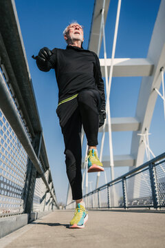 A fit senior man performs high knee warm-up exercises while jogging across a contemporary white bridge under a clear blue sky, focusing on his health and active lifestyle.