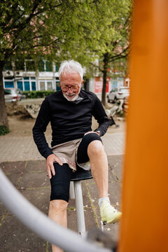 Active senior man with white hair wearing compression knee sleeves while training on stationary bicycle equipment at an outdoor public gym.