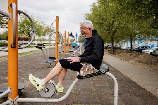 Active senior man with a beard and glasses sits on an outdoor fitness machine while pedaling a stationary bike at a public park workout station.