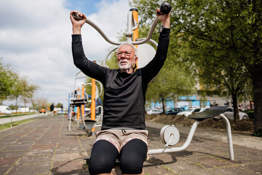 Senior man focuses on his fitness while using a shoulder press machine at an outdoor community gym in a public park during his workout routine.