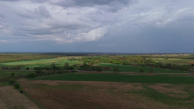 Aerial orbit over farmland with patchy sunlight breaking through clouds across fields and trees.