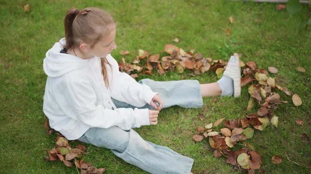 Young girl crafts outdoors creatively. Child meticulously arranges leaves into circle during play. Young girl with ponytail braid intricately arranges leaves into artistic circle outside