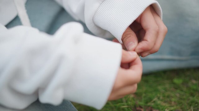 Close up hands in white hoodie small child fingers fidgeting with sleeve while seated on grass soft natural light cozy casual clothing intimate candid moment conveying shy introspection and subtle